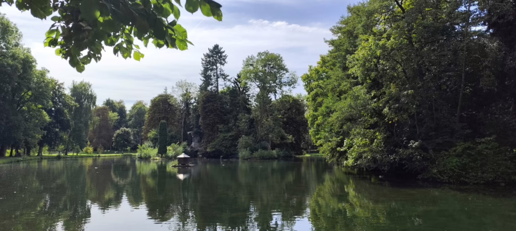 Gartenlandschaft in Donaueschingen, blauer Himmel mit hellen Wolken, ein großer See und am Ufer üppige Bäume.