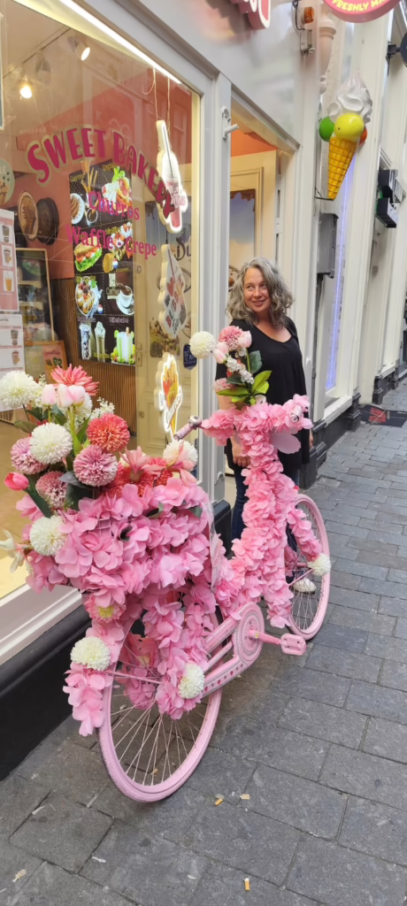 Frances vor rosa geschmmücktem Fahrrad vor einem niederländischen Bäcker. Blick zurück ins Ladengeschäft. Während unseres Besuches in Amsterdam 2025