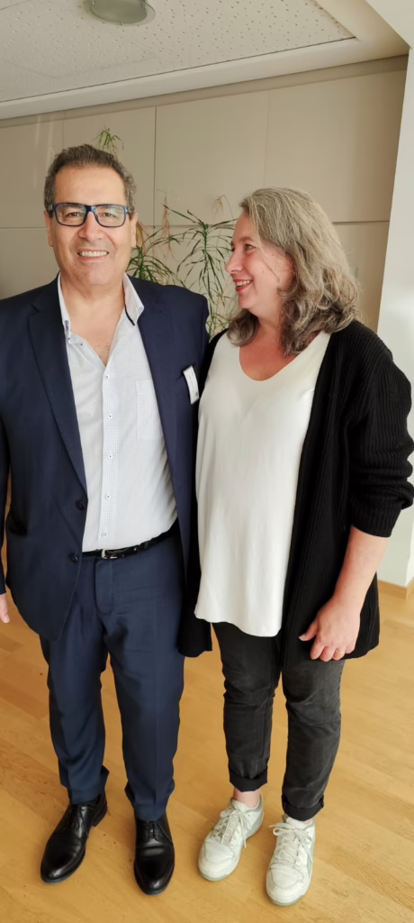 Dr. Ferdosi, Chefarzti der Senologie im Anzug und weißen Hemd und frances mit weißem T-shirt und dunkler Strickjacke. Ferdosi schaut genau in die Kamera, Frances schaut zu ihrem Chirurgen und strahlt.